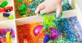 Sensory box with rainbow rice inside. Children's hands touch the texture and pour. Tool for sorting tongs and spoon. Montessori material baby
